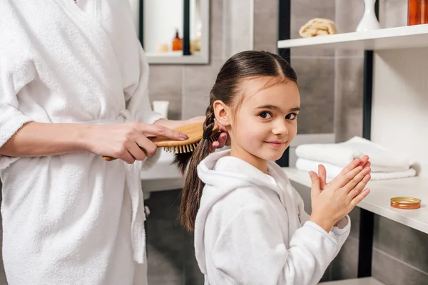 Mother in white bathrobe combing daughter hair with wooden hairbrush near shelves in bathroom — Stock Photo