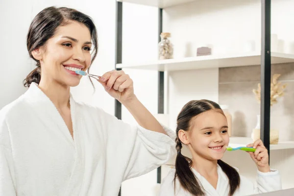 Mother and daughter in white bathrobes brushing teeth in bathroom — Stock Photo