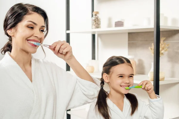 Mother and daughter in white bathrobes brushing teeth with toothbrushes in bathroom — Stock Photo