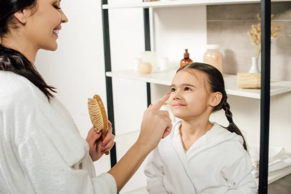 Madre sosteniendo cepillo de pelo y tocando la nariz hija en el baño - foto de stock