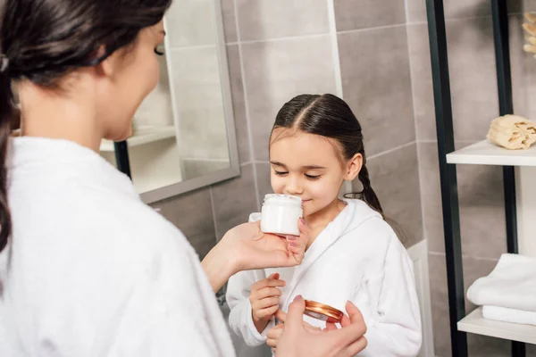 Daughter sniffing cosmetic cream holding mother in bathroom — Stock Photo