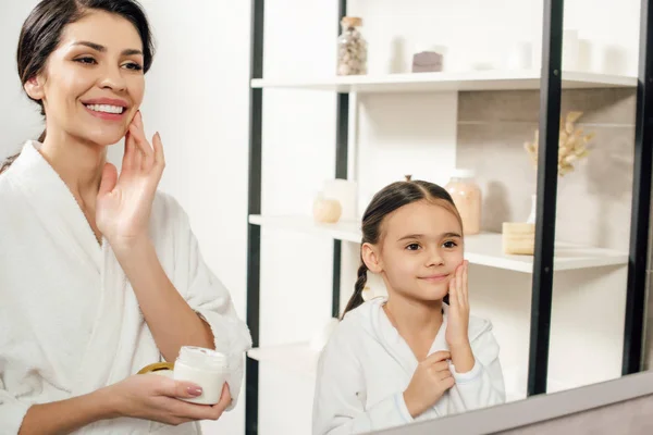 Мать и дочь в белых халатах наносят косметический крем в ванной комнате — стоковое фото
