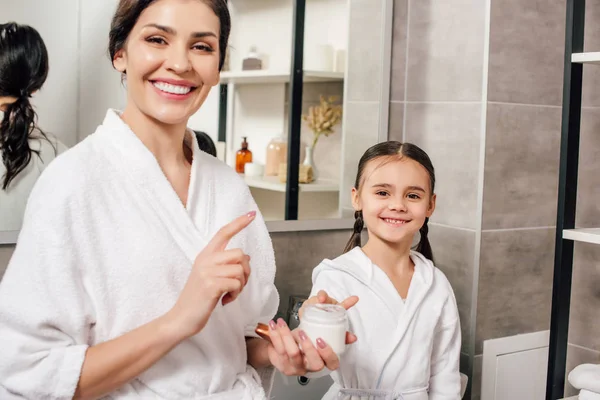 Madre e hija en batas blancas con crema cosmética en el baño - foto de stock