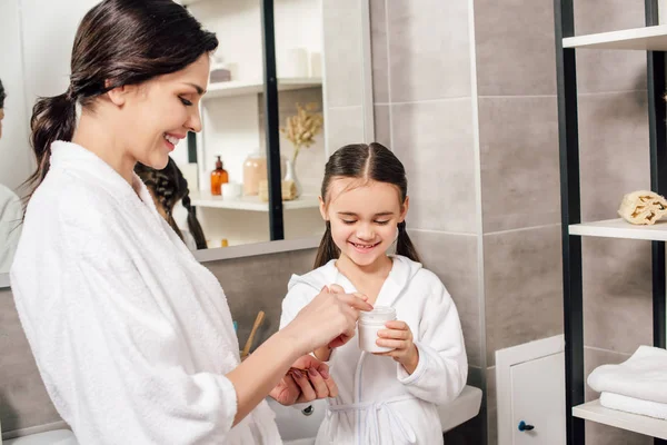 Mother and daughter in white bathrobes with container of cosmetic cream in bathroom — Stock Photo