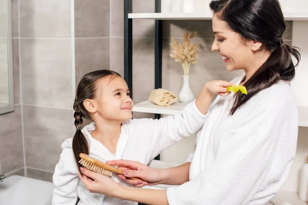 Mother and daughter in white bathrobes combing each other in bathroom — Stock Photo