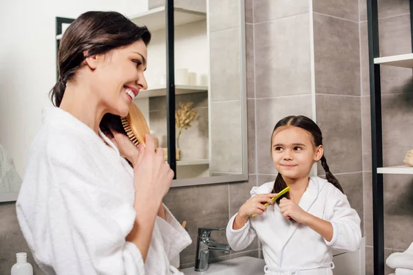 Mother and daughter in white bathrobes combing in bathroom — Stock Photo