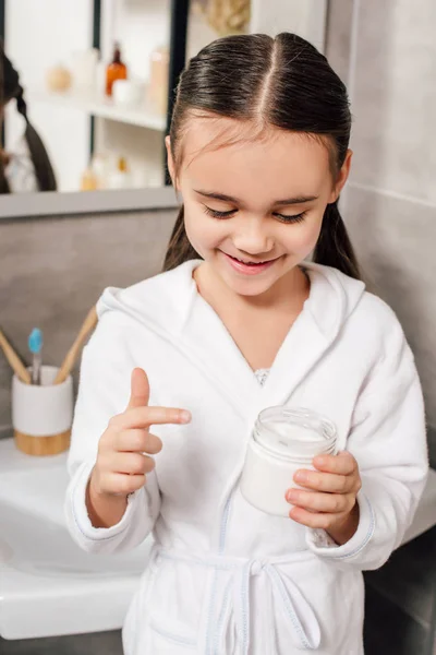 Child in white bathrobe holding container with cosmetic cream in bathroom — Stock Photo