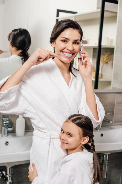 Daughter hugging mother brushing teeth and talking on smartphone in bathroom — Stock Photo