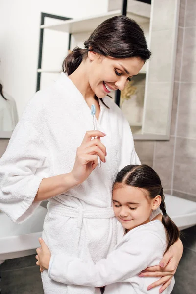 Дочь обнимает мать держа зубную щетку на смартфоне в ванной комнате — стоковое фото