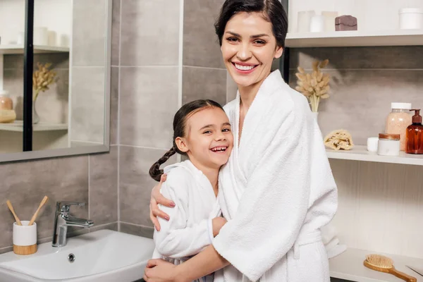 Hija y madre en batas blancas abrazándose y sonriendo en el baño - foto de stock