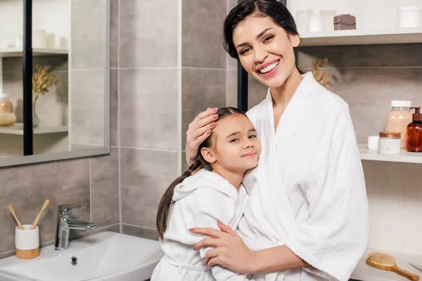 Дочь и мать в белых халатах обнимаются в ванной — стоковое фото