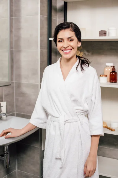 Frau im weißen Bademantel steht und lächelt im Badezimmer — Stockfoto