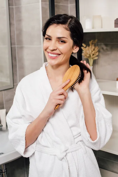 Donna in accappatoio bianco pettinando i capelli e sorridendo in bagno — Foto stock