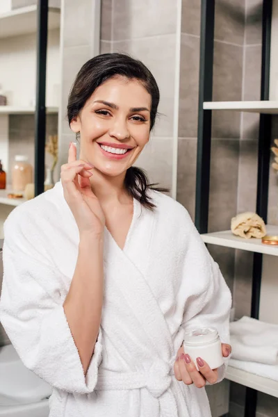 Frau im Bademantel hält Behälter und trägt Kosmetikcreme im Badezimmer auf — Stockfoto