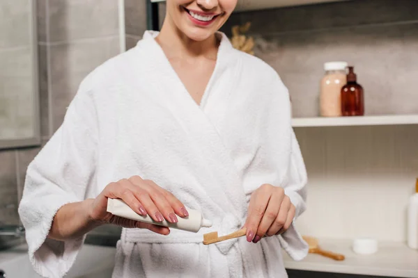 Обрезанный вид женщины, наносящей зубную пасту на зубную щетку из тюбика в ванной комнате — стоковое фото