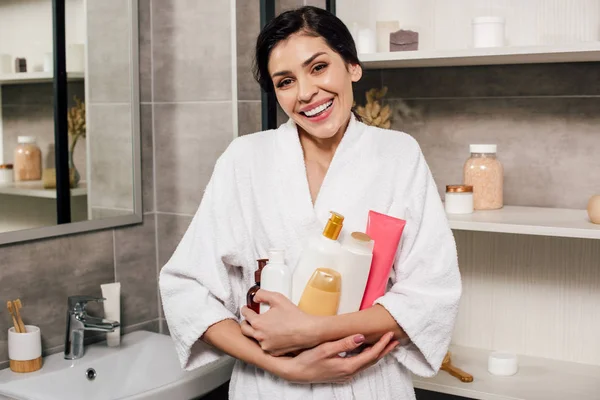 Woman in white bathrobe holding bottles and smiling in bathroom — Stock Photo