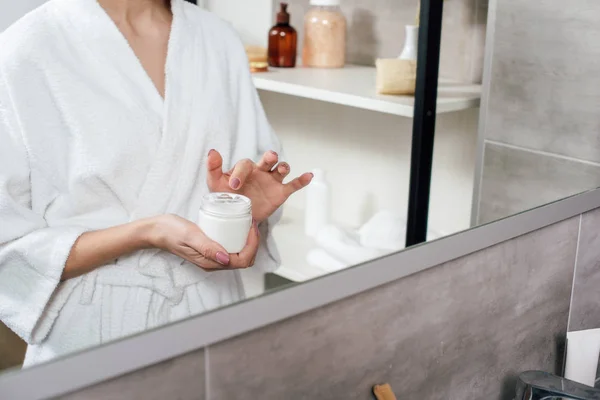 Cropped view of woman in white bathrobe holding container with cosmetic cream in bathroom — Stock Photo