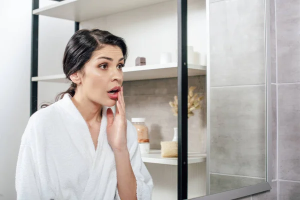 Agitated woman in white bathrobe looking to mirror in bathroom — Stock Photo