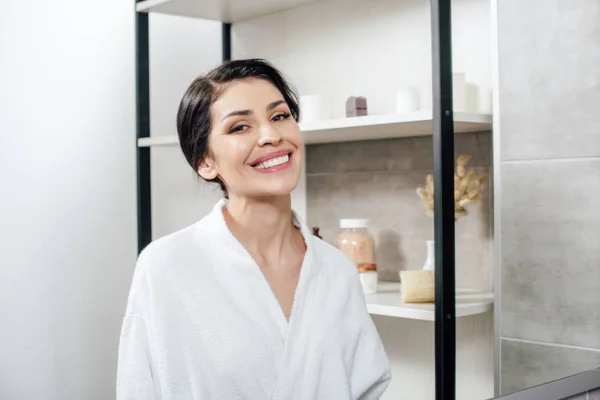Женщина в белом халате смотрит в зеркало и улыбается в ванной комнате — стоковое фото