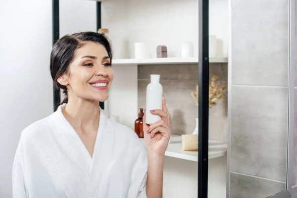 Woman in white bathrobe looking to mirror, holding bottle with lotion and smiling in bathroom — Stock Photo