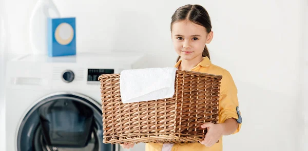 Panoramic shot of child standing and holding basket with towel in laundry room — Stock Photo