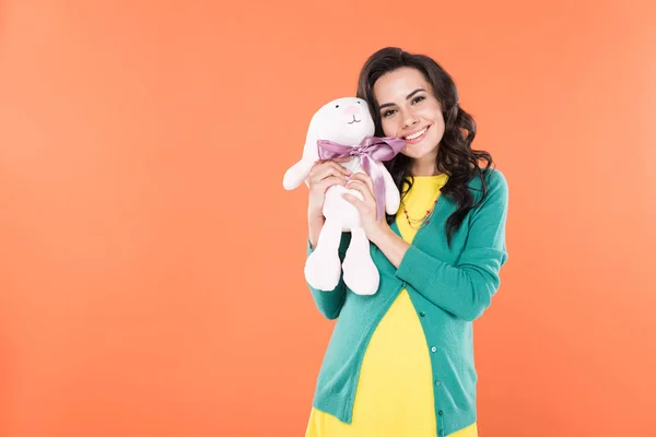 Чарівна брюнетка вагітна жінка тримає іграшкового кролика і посміхається ізольовано на помаранчевому — стокове фото