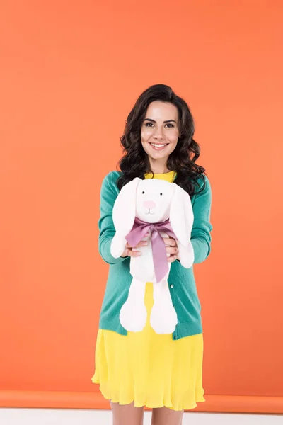 Vue de face de joyeuse femme enceinte tenant lapin jouet avec sourire sur fond orange — Photo de stock