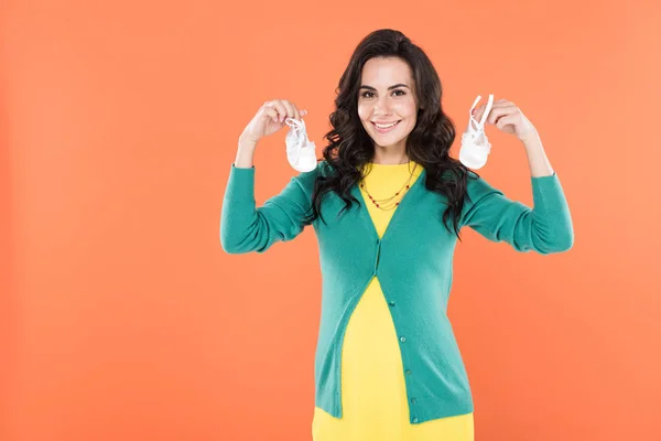 Attractive brunette pregnant woman holding baby shoes isolated on orange — Stock Photo