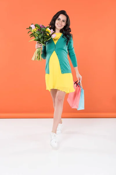 Vista completa de la mujer embarazada sonriente sosteniendo flores y bolsas de compras sobre fondo naranja - foto de stock
