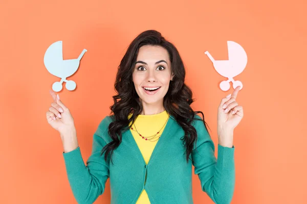 Amazed brunette woman in green cardigan pointing with fingers at paper baby carriages on orange background — Stock Photo