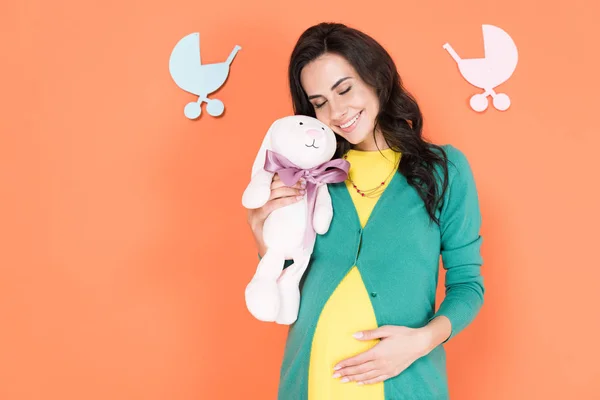 Hermosa mujer embarazada sonriendo con los ojos cerrados y sosteniendo el conejo de juguete sobre fondo naranja - foto de stock