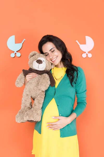 Mujer embarazada feliz sosteniendo oso de peluche y tocando el vientre con los ojos cerrados sobre fondo naranja - foto de stock