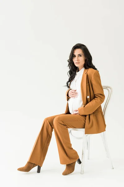 Беременная женщина в коричневом костюме сидит на стуле на белом фоне — стоковое фото