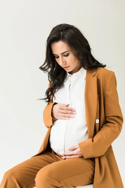 Брюнетка беременная женщина в коричневом костюме касается живота и смотрит вниз изолированы на белый — стоковое фото