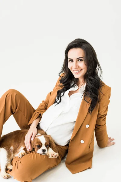 Preciosa mujer embarazada en traje marrón acariciando perro sobre fondo blanco - foto de stock