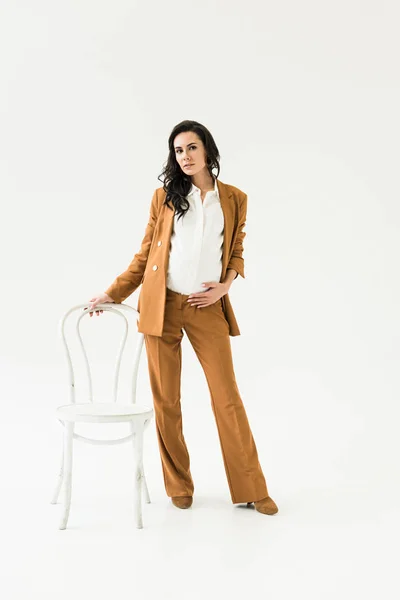 Stylish pregnant woman in suit standing near chair on white background — Stock Photo