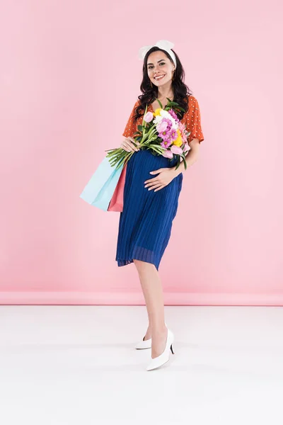 Mujer embarazada en falda azul sosteniendo flores y bolsas de compras sobre fondo rosa - foto de stock