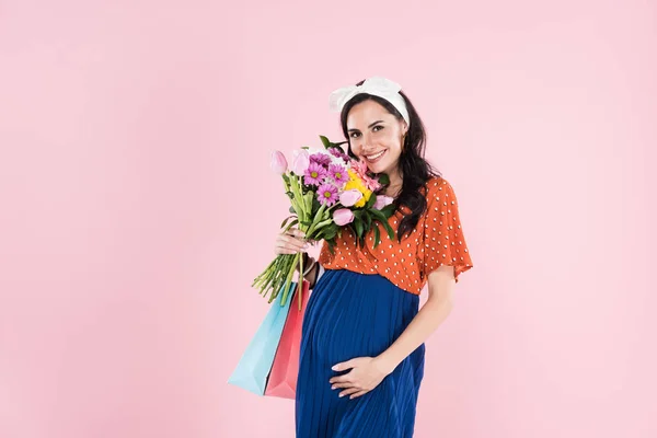 Femme enceinte romantique tenant des sacs à provisions et des fleurs isolées sur rose — Photo de stock