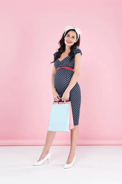 Stylish pregnant woman in dotted dress holding shopping bags on pink background — Stock Photo