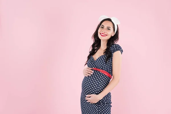 Mujer embarazada sonriente en vestido punteado tocando vientre aislado en rosa - foto de stock