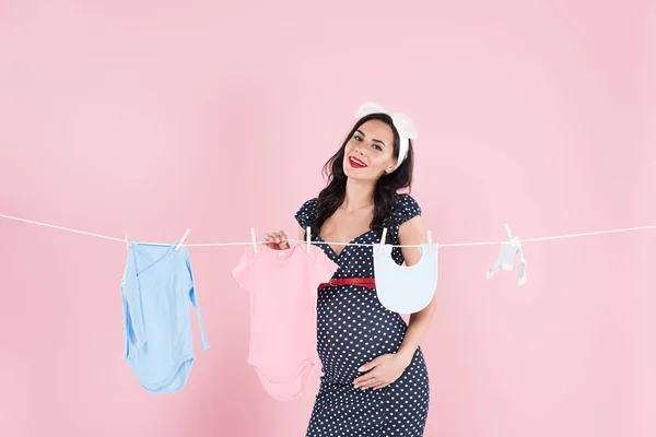 Pregnant woman in dotted dress hanging out baby clothes on clothesline on pink background — Stock Photo