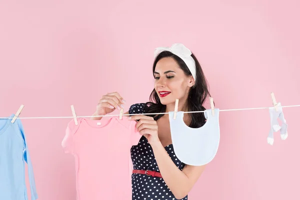 Charming pregnant woman hanging out baby clothes on clothesline isolated on pink — Stock Photo