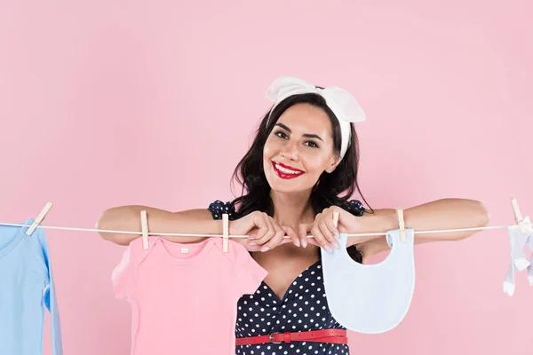 Jolie brune jeune femme accrochant des vêtements de bébé sur corde à linge isolé sur rose — Photo de stock