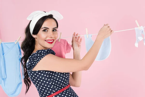 Affascinante donna incinta appendere fuori vestiti bambino con sorriso isolato su rosa — Foto stock