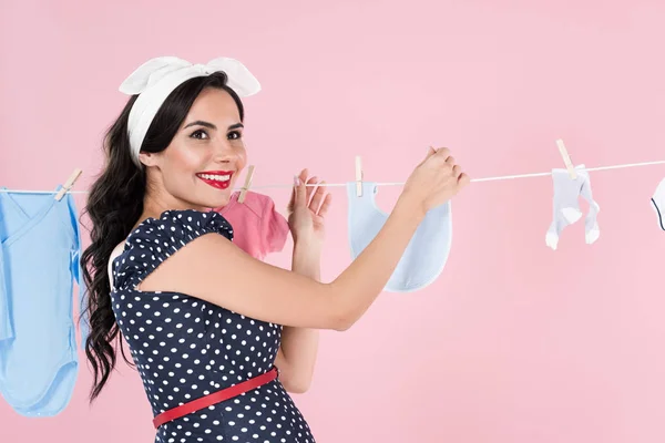 Excited pregnant woman hanging out baby clothes on clothesline isolated on pink — Stock Photo