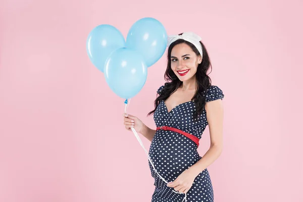 Mujer embarazada feliz en vestido de lunares sosteniendo globos de aire azul aislados en rosa - foto de stock