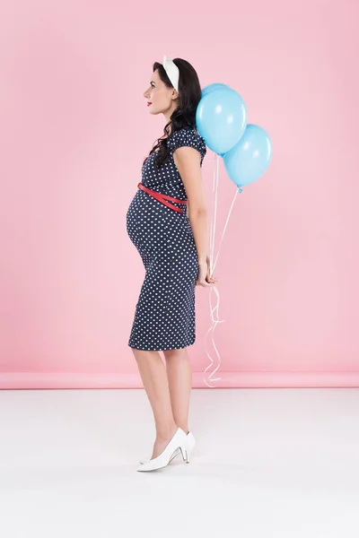 Side view of pregnant woman in dotted dress holding air balloons on pink background — Stock Photo