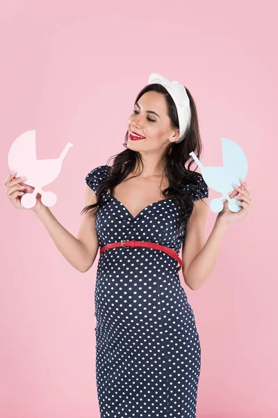 Happy brunette pregnant woman holding paper strollers isolated on pink — Stock Photo