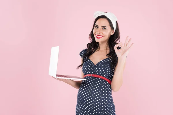 Mujer embarazada feliz sosteniendo el ordenador portátil y mostrando signo bien aislado en rosa - foto de stock