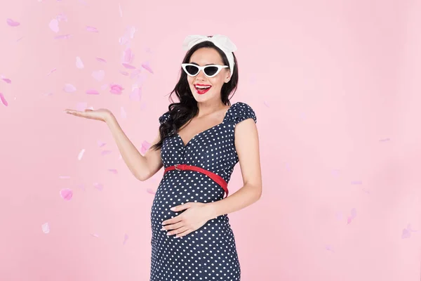 Веселая беременная женщина в солнечных очках, стоящая под конфетти изолированы на розовый — стоковое фото